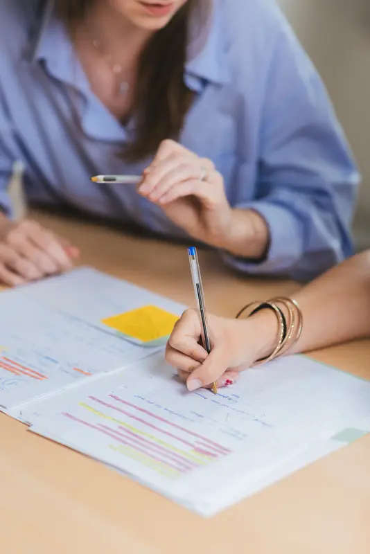 Gros plan sur la main d'une élève annotant son cahier, la fondatrice en arrière-plan